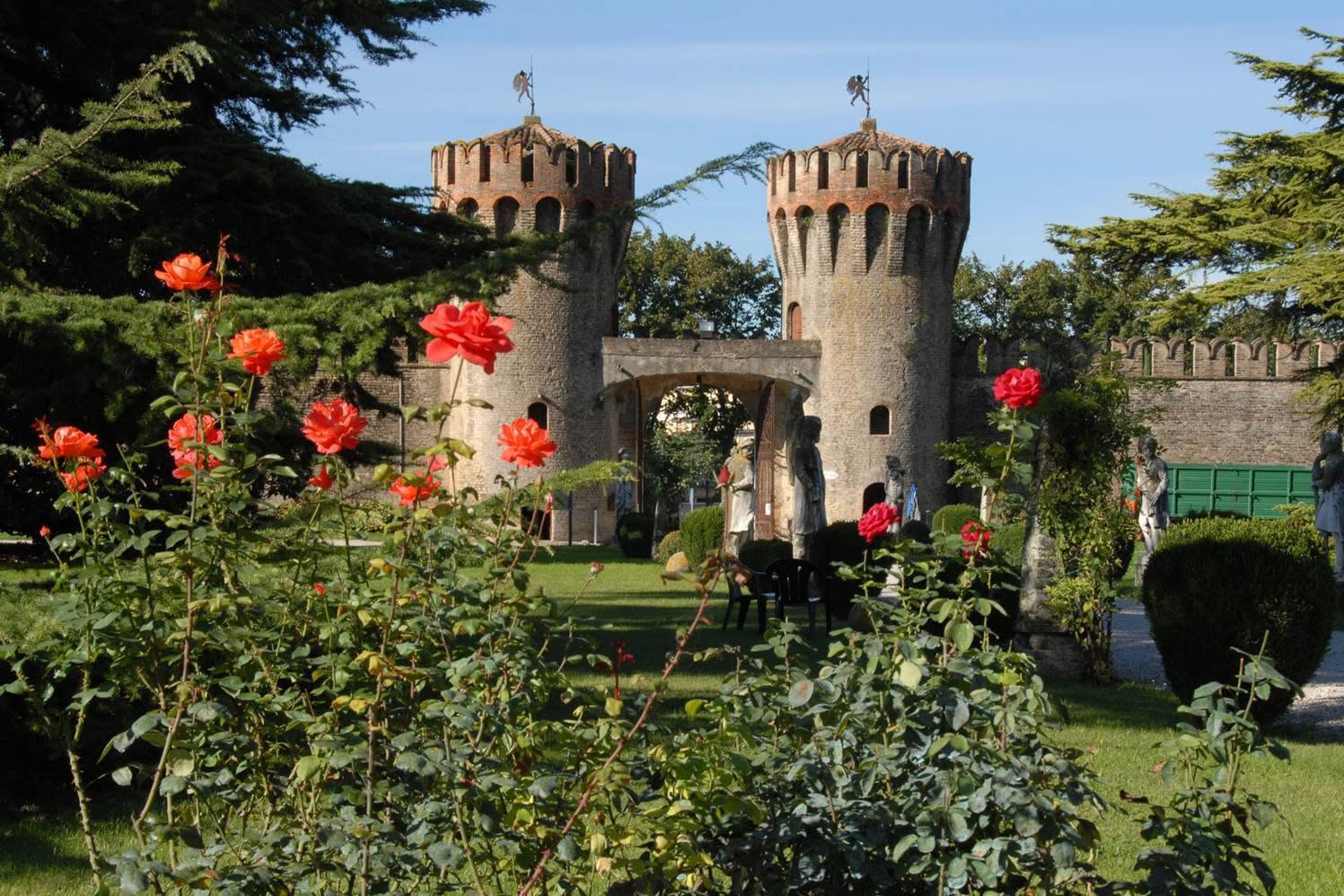 A stone castle entrance with two round towers stands behind a rose garden in clear daylight.